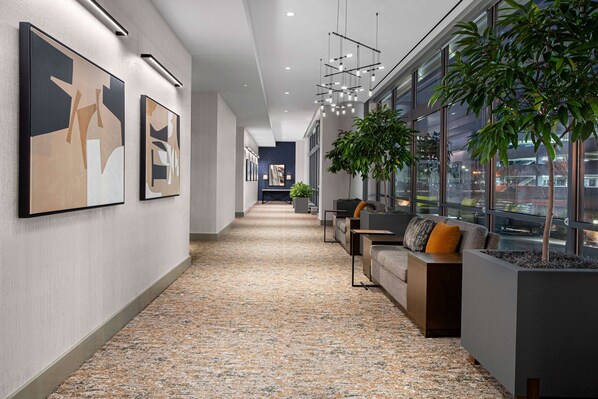 A modern hallway with abstract art, a seating area with a sofa and a coffee table, and a large window with a view of the outside. - Hilton Garden Inn Camden Waterfront Philadelphia (Camden)