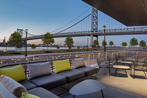 An outdoor seating area with a view of a bridge, tables, and chairs. - Hilton Garden Inn Camden Waterfront Philadelphia (Camden)