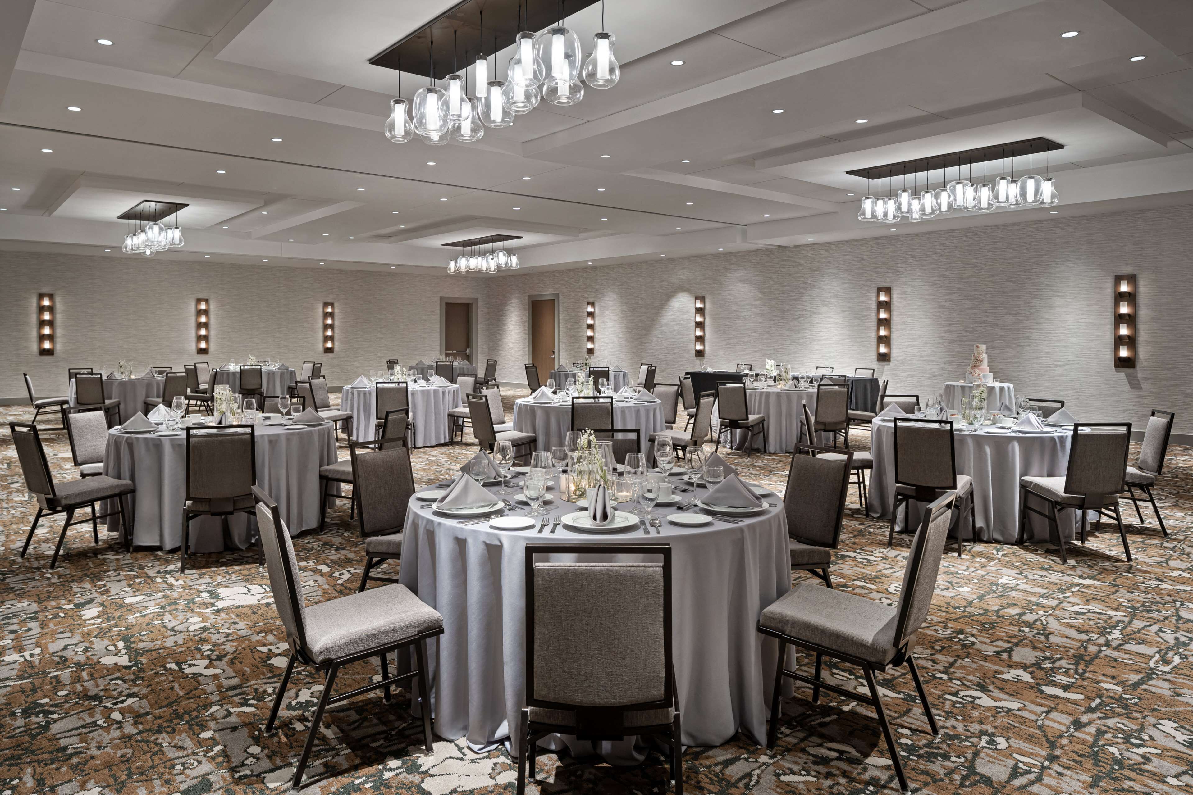 A large banquet hall with round tables set for dining, featuring a patterned carpet and modern lighting fixtures.
