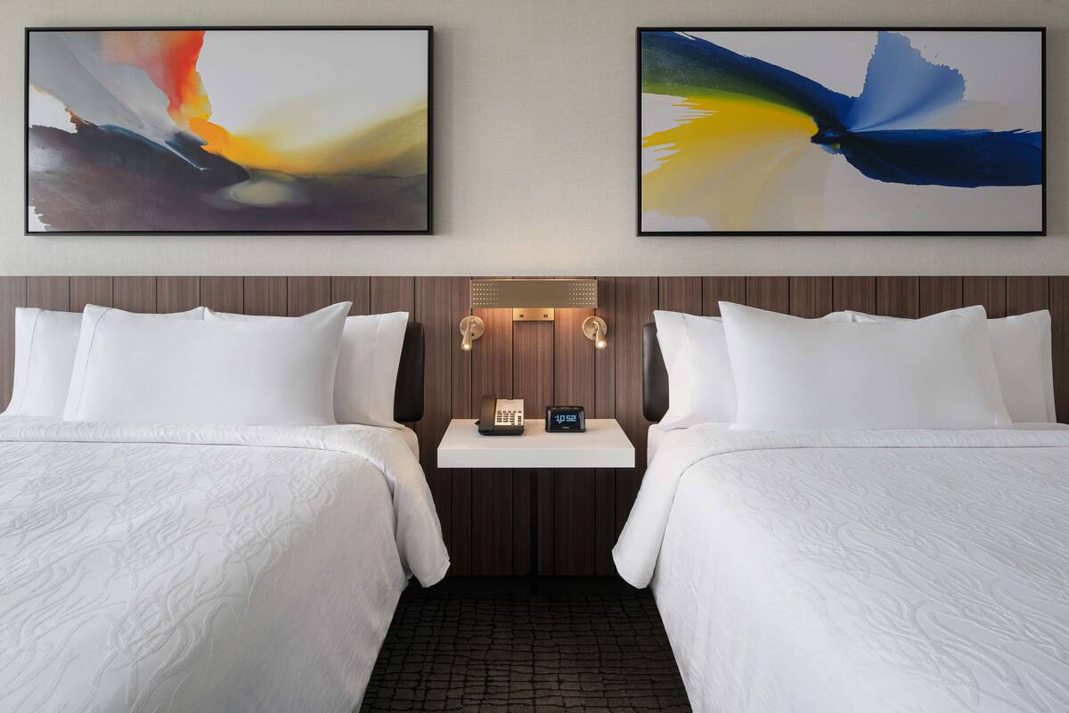 A hotel room with two beds, a small table with a phone and clock, and abstract paintings on the wall.