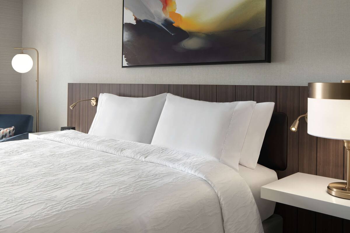 A neatly made bed with white linens, a modern bedside table with a lamp, and a large abstract painting on the wall.