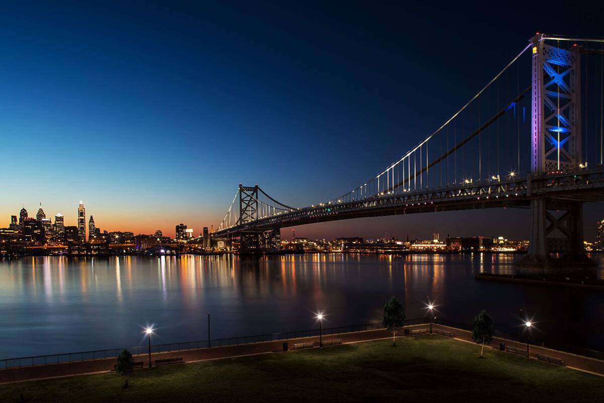 A nighttime view of a large suspension bridge spanning a wide river, with city lights reflecting on the water.