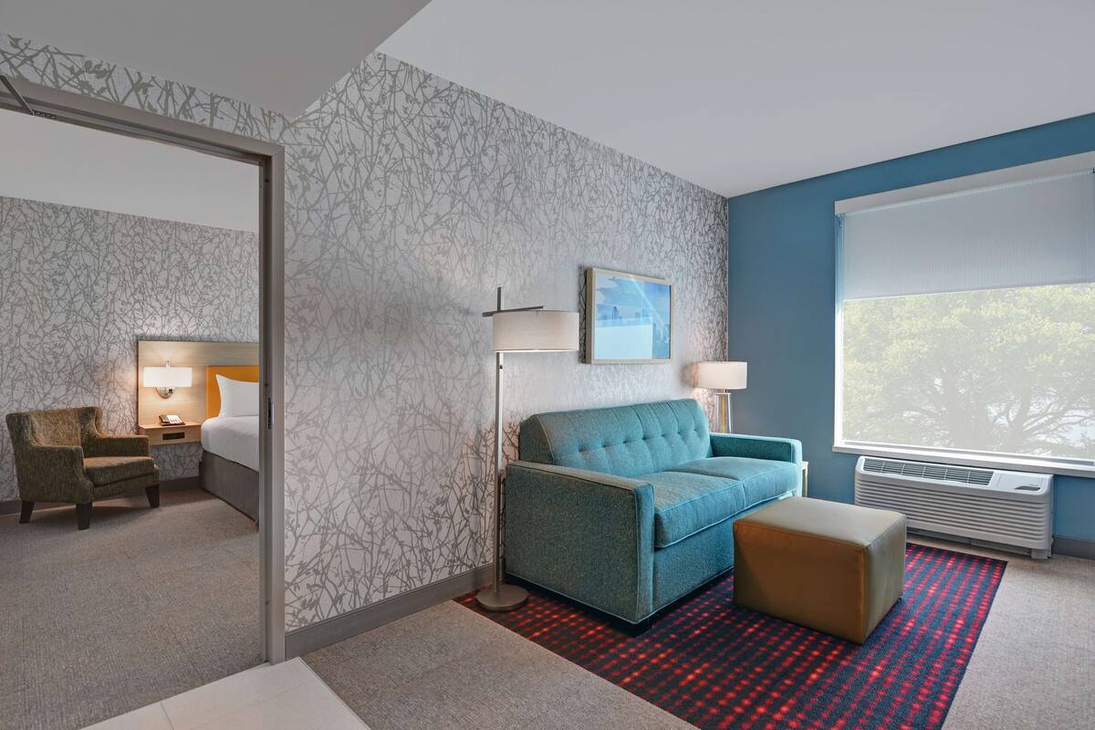Une chambre d’hôtel avec un canapé bleu, un petit pouf, une chaise, une table de chevet avec une lampe, un tableau encadré au mur et une grande fenêtre donnant sur des arbres.