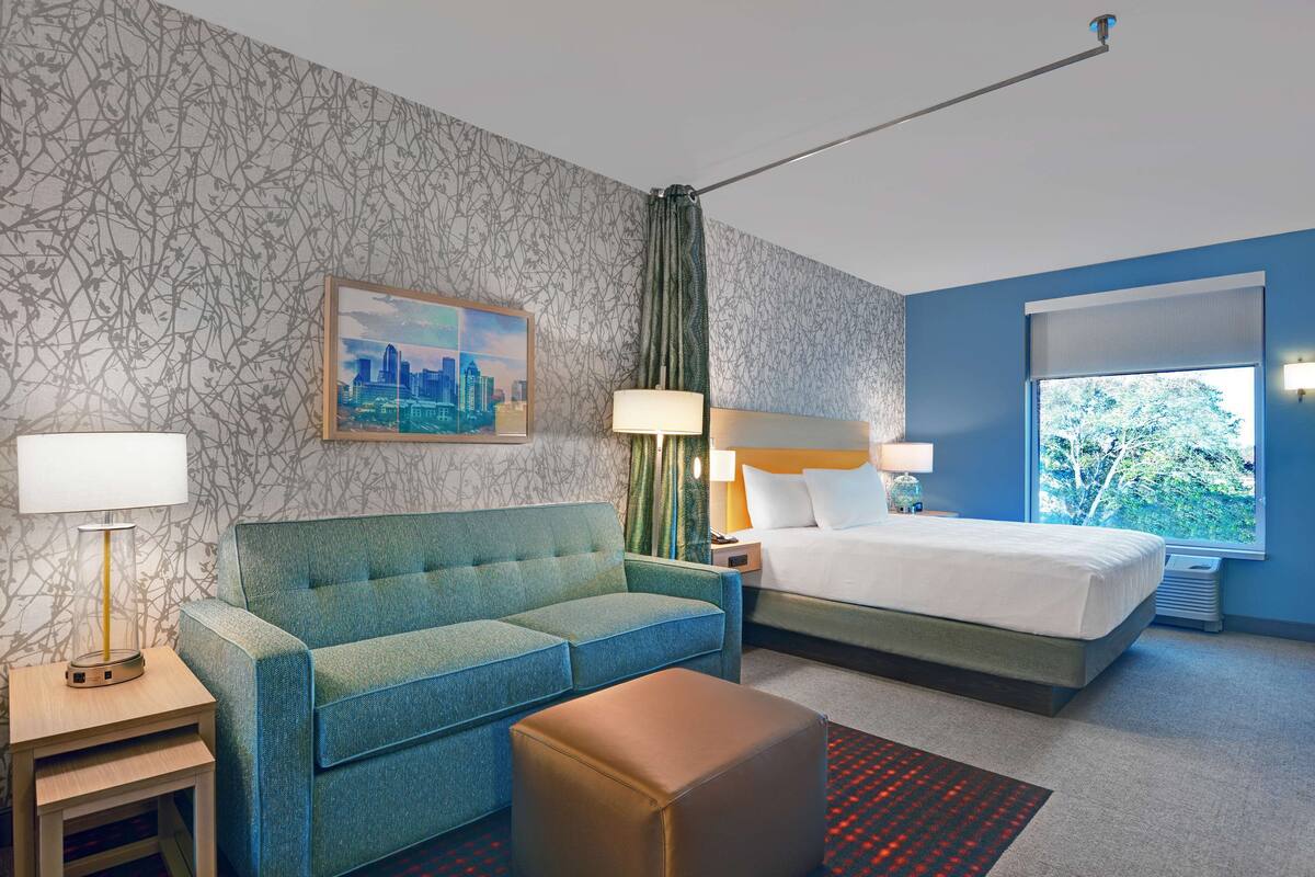 Une chambre d’hôtel avec un lit, un canapé, une table de chevet, une lampe, un tableau et une fenêtre donnant sur un paysage.