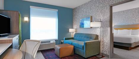 A hotel room with a blue wall, a large window, a sofa, a TV, a desk, and a bed.