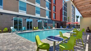 A pool area with a brick floor, green chairs, and tables, surrounded by a building with large windows and a red section. - Home2 Suites by Hilton Largo (Largo)