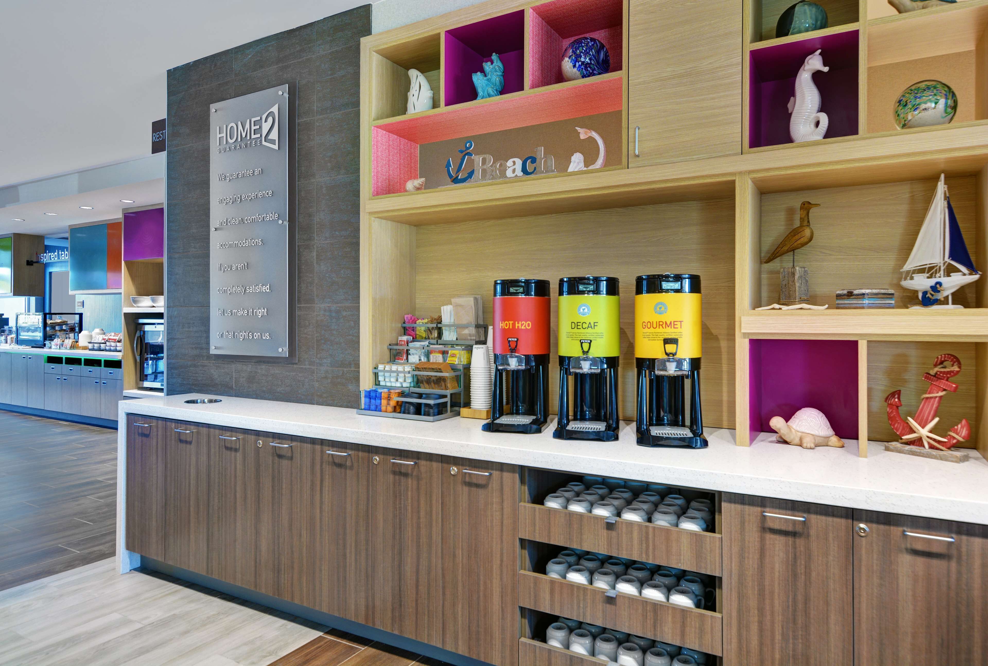 A hotel reception area with a coffee station, a sign for 'Home2 Guarantee', and various decorative items on shelves.