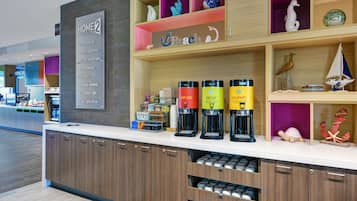 Een hotel receptie met een koffiehoek, een bord met 'Home2 Guarantee' en diverse decoratieve objecten op planken.