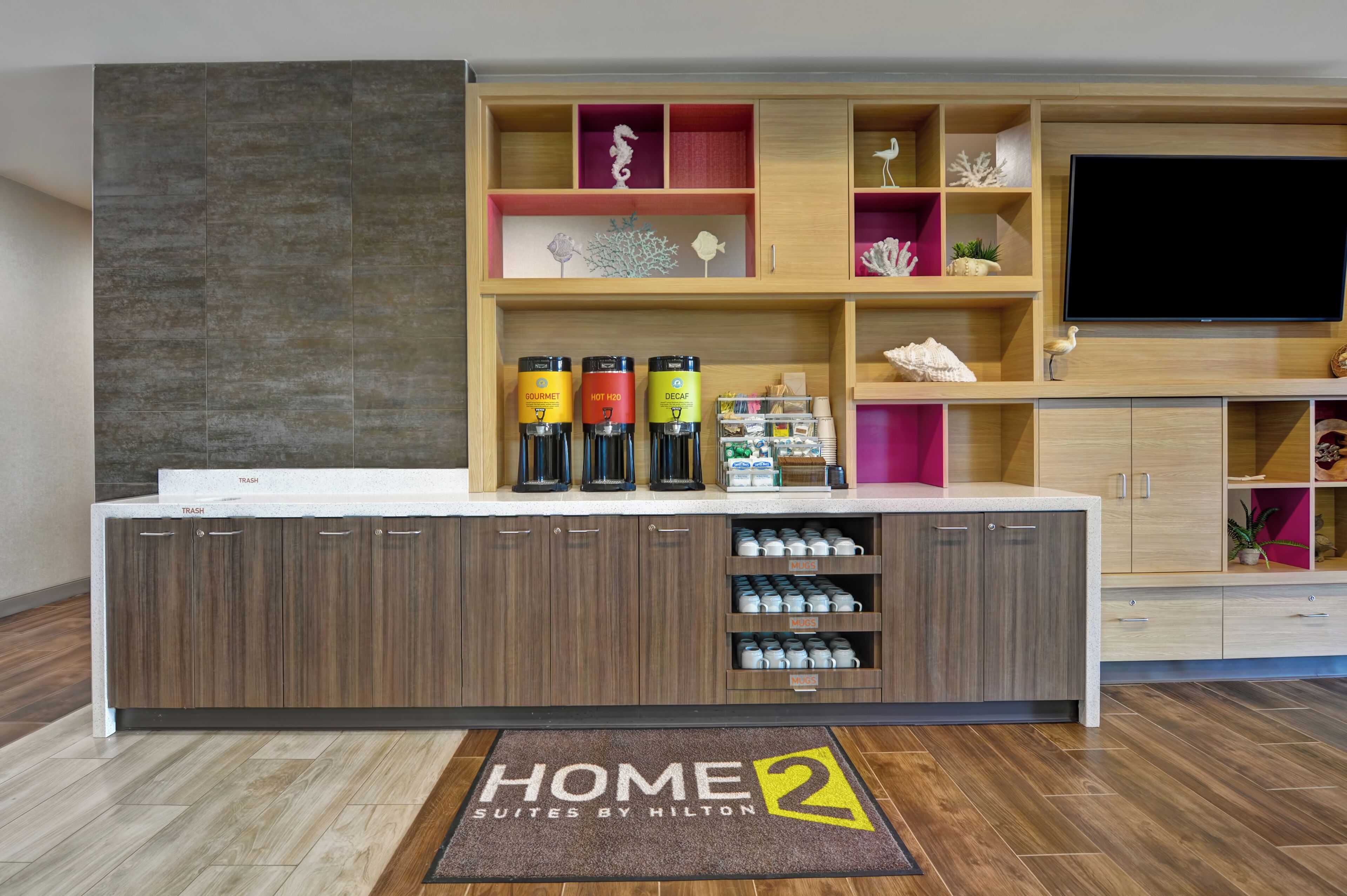 A hotel reception area with a counter, shelves with decorative items, a television, and a 'HOME 2 SUITES BY HILTON' mat.