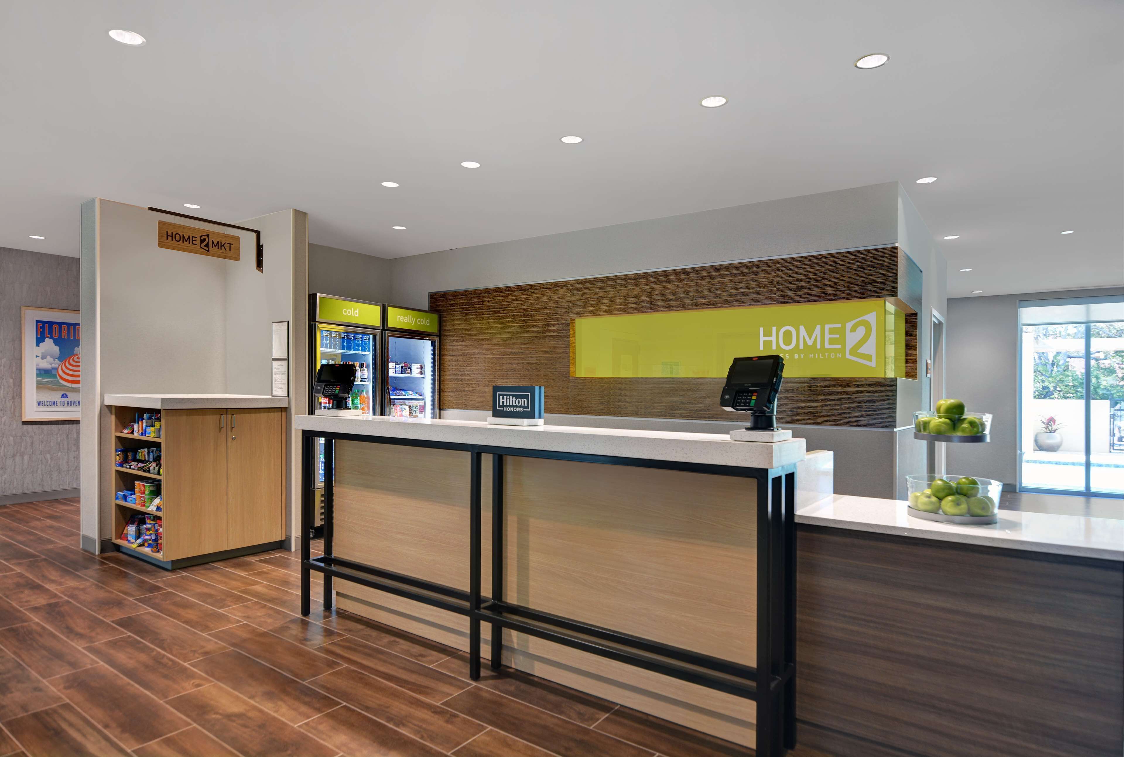 Un espace d’accueil moderne, avec un comptoir en bois, un écran numérique affichant le mot « HOME2 » et un distributeur de boissons en libre-service.