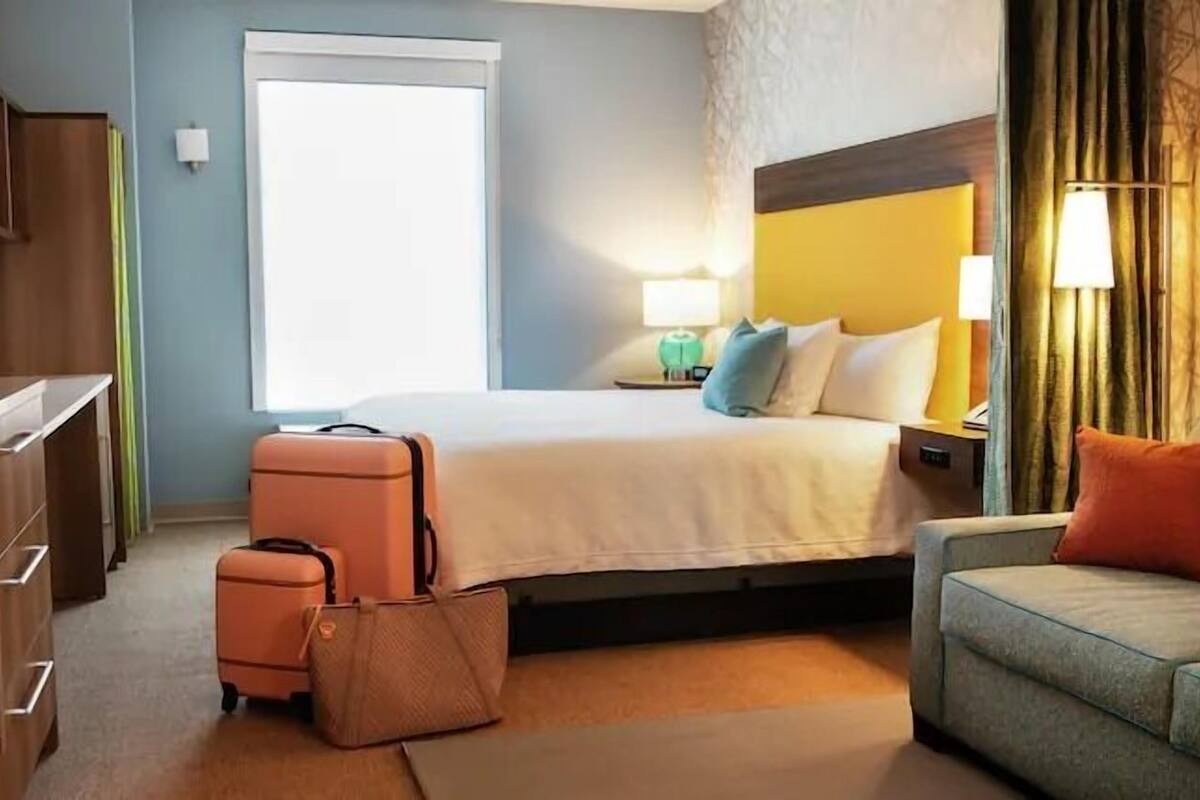 Une chambre d’hôtel avec un lit, un bureau, une chaise, une valise et une petite table avec une lampe.