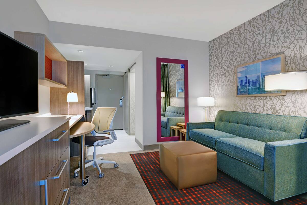 Une chambre d’hôtel moderne avec un bureau, un canapé bleu, un petit pouf, une télévision et un tableau représentant un paysage urbain accroché au mur.