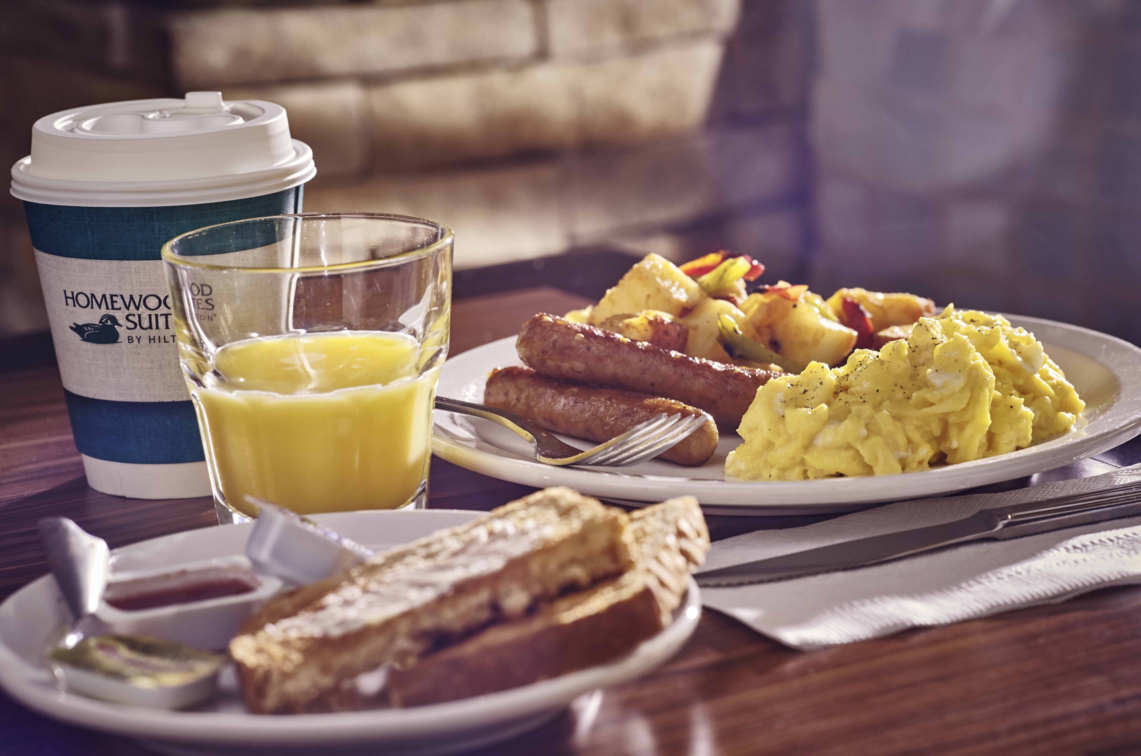 Une table dressée pour le petit-déjeuner avec une tasse de café, un verre de jus d’orange, des toasts, des œufs brouillés, des saucisses et des pommes de terre rôties.