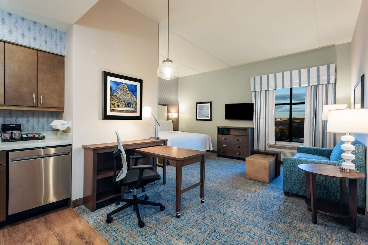 A hotel room with a kitchenette, a bed, a desk, a chair, a sofa, and a TV.