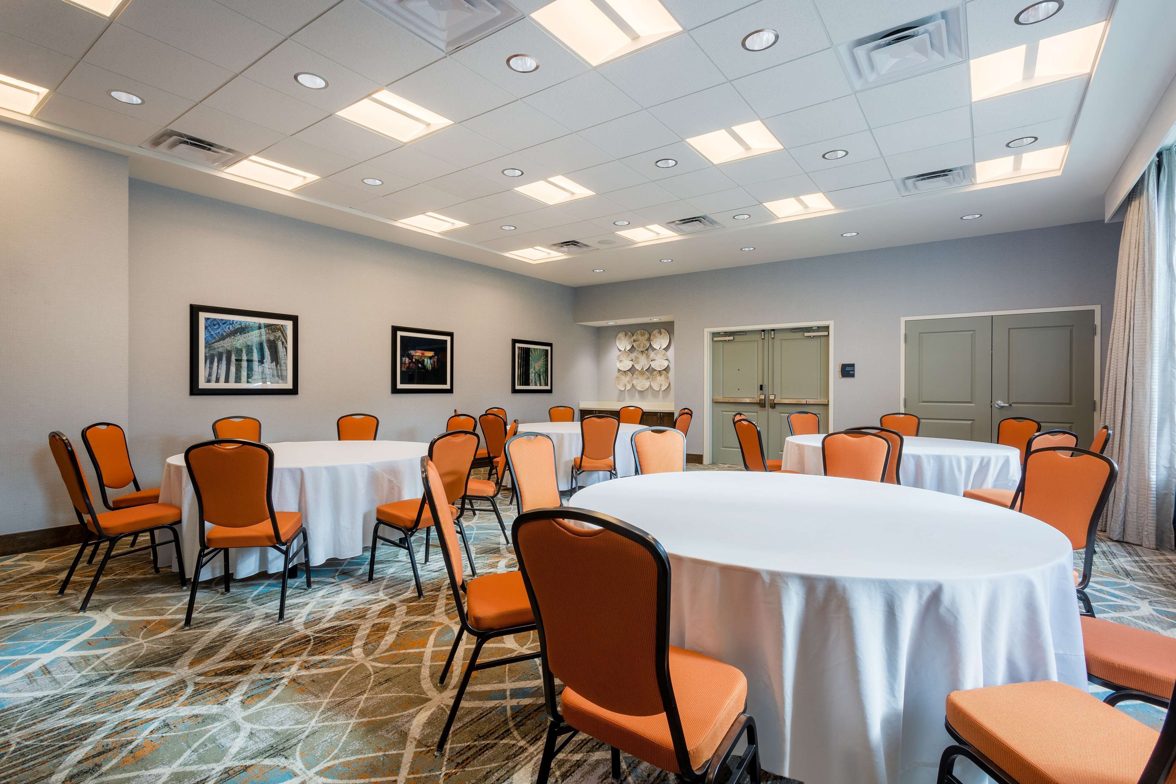 A conference room with multiple round tables and orange chairs, white tablecloths, and a patterned carpet.