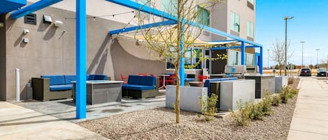 An outdoor seating area with blue and gray furniture, a small tree, and a building with large windows in the background.