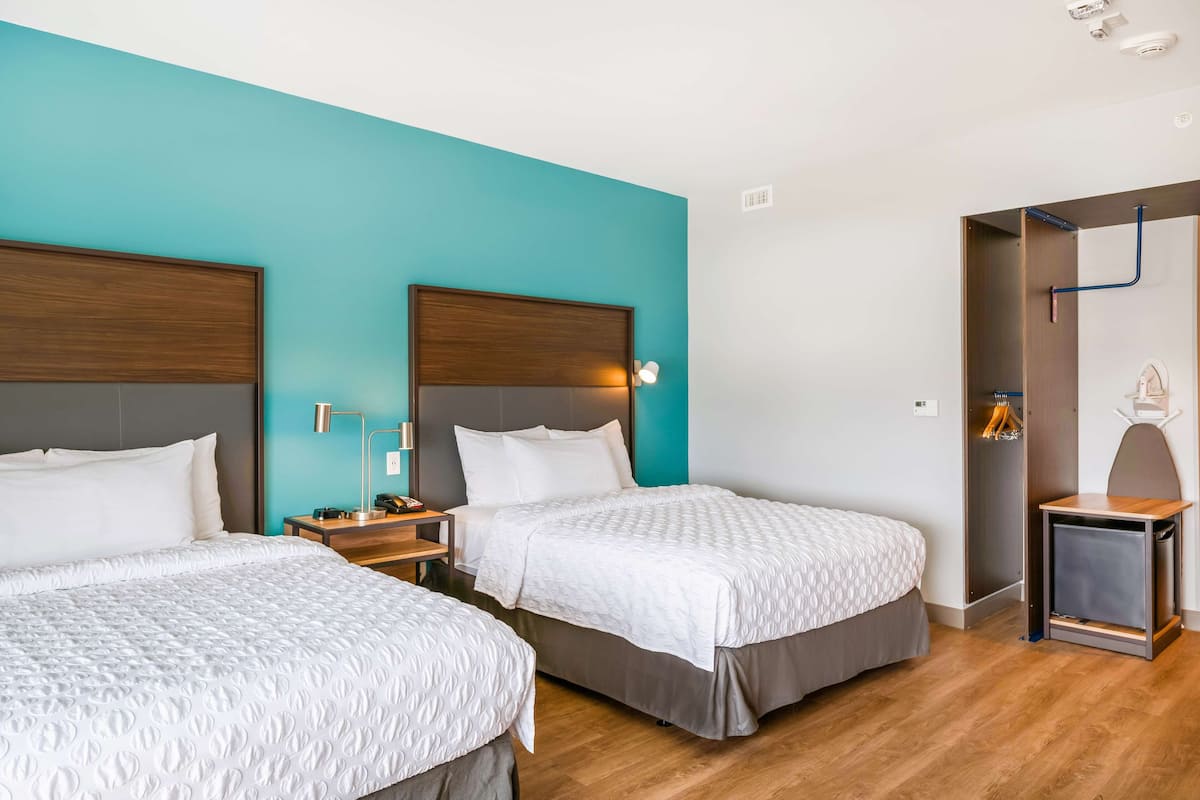 A hotel room with two beds, a wooden floor, a teal accent wall, and a small seating area with a table and chair.