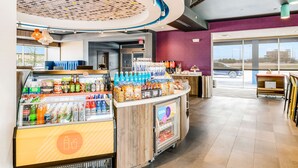 Un moderno quiosco de bebidas con diversas opciones de bebidas y aperitivos, un área de estar con una pared morada y un amplio ventanal con vistas al exterior.