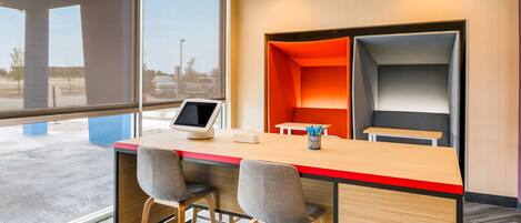 A modern office space with a large window, a desk with two chairs, and a seating area with orange and grey cushions.