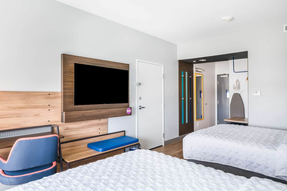A hotel room with two beds, a TV mounted on the wall, a desk with a chair, and a bathroom visible through an open door.