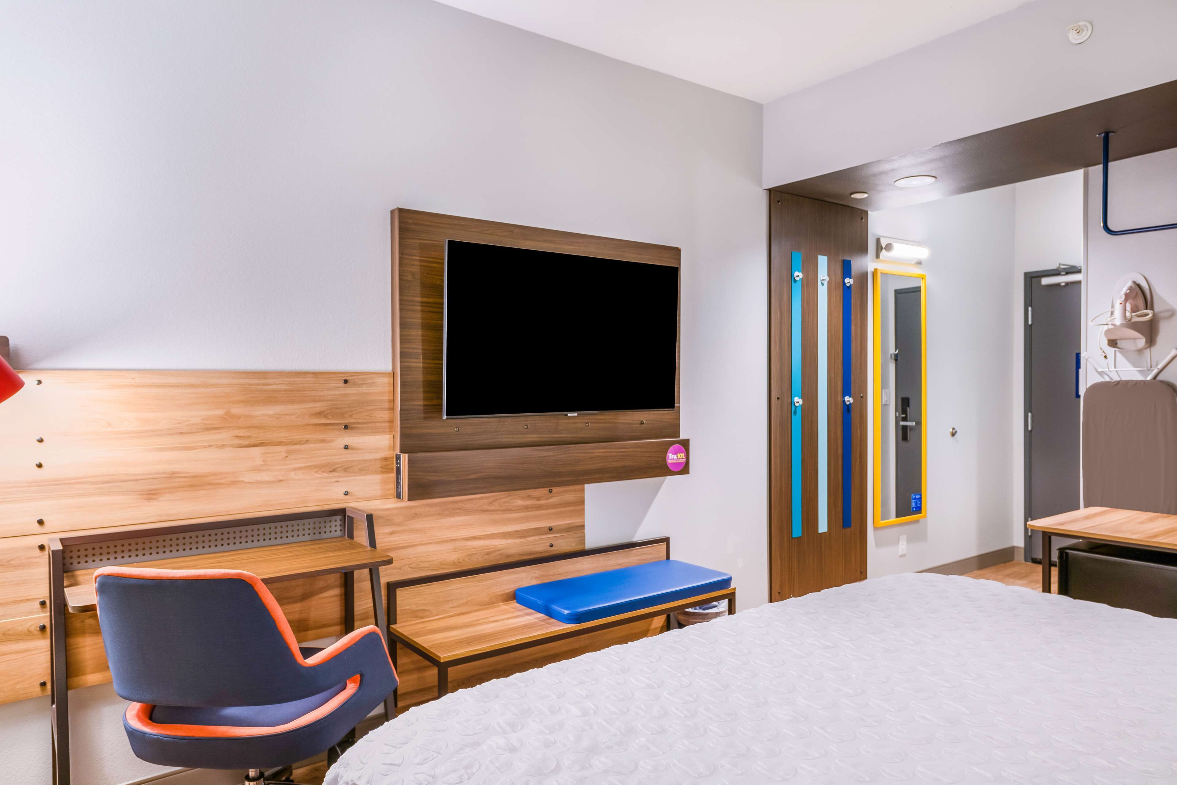 Quarto de hotel com uma cama, uma escrivaninha com cadeira, uma televisão na parede e um espelho com listras verticais.