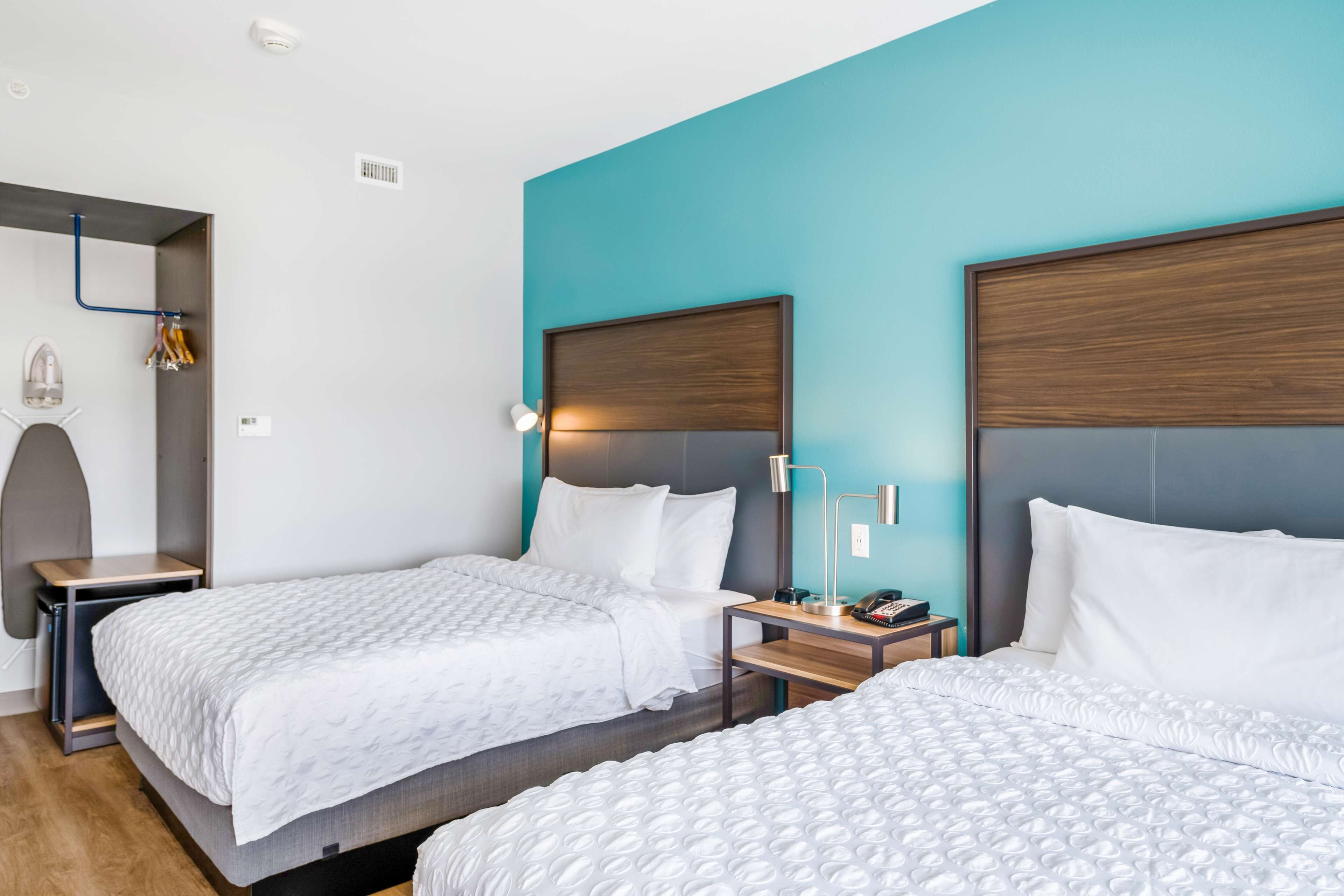 A hotel room with two beds, a blue accent wall, a wooden headboard, a bedside table with a lamp, and a small shelf with decorative items.
