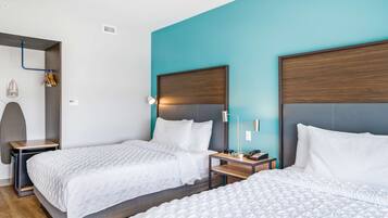 A hotel room with two beds, a blue accent wall, a wooden headboard, a bedside table with a lamp, and a small shelf with decorative items.