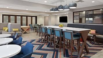 A modern hotel lobby with a long communal table, several bar stools, a sofa, and a small round table.