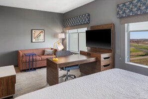 A modern hotel room with a large bed, a desk with a chair, a sofa, a TV, and a window with a view of the countryside.