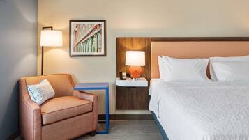Habitación de hotel con una cama, una silla, una mesita de noche con lámpara y un cuadro en la pared.