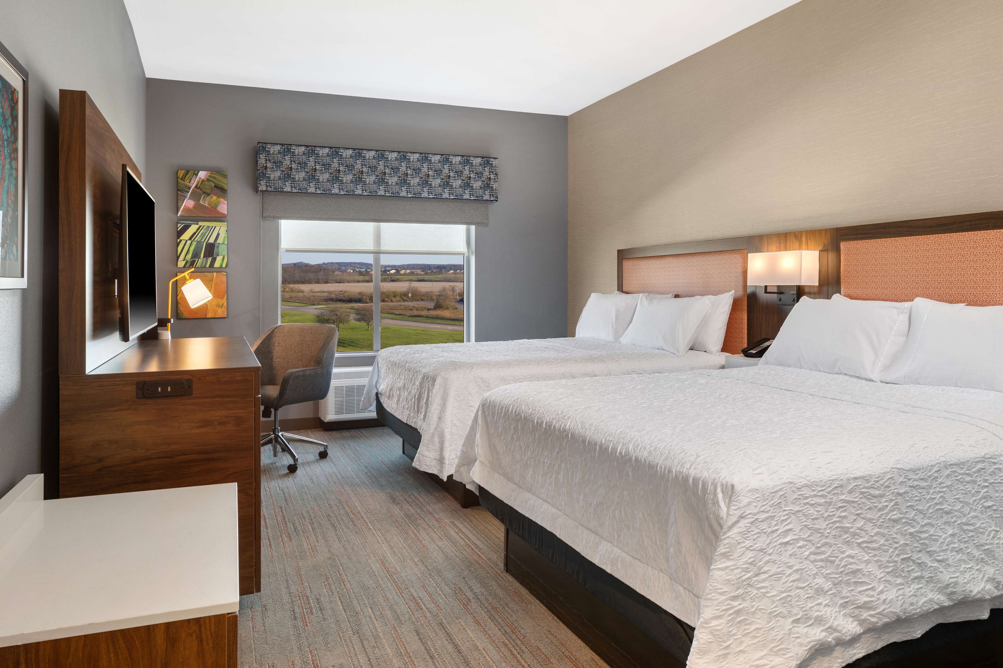 Habitación de hotel con dos camas, un escritorio, una silla y un ventanal con vistas al campo.