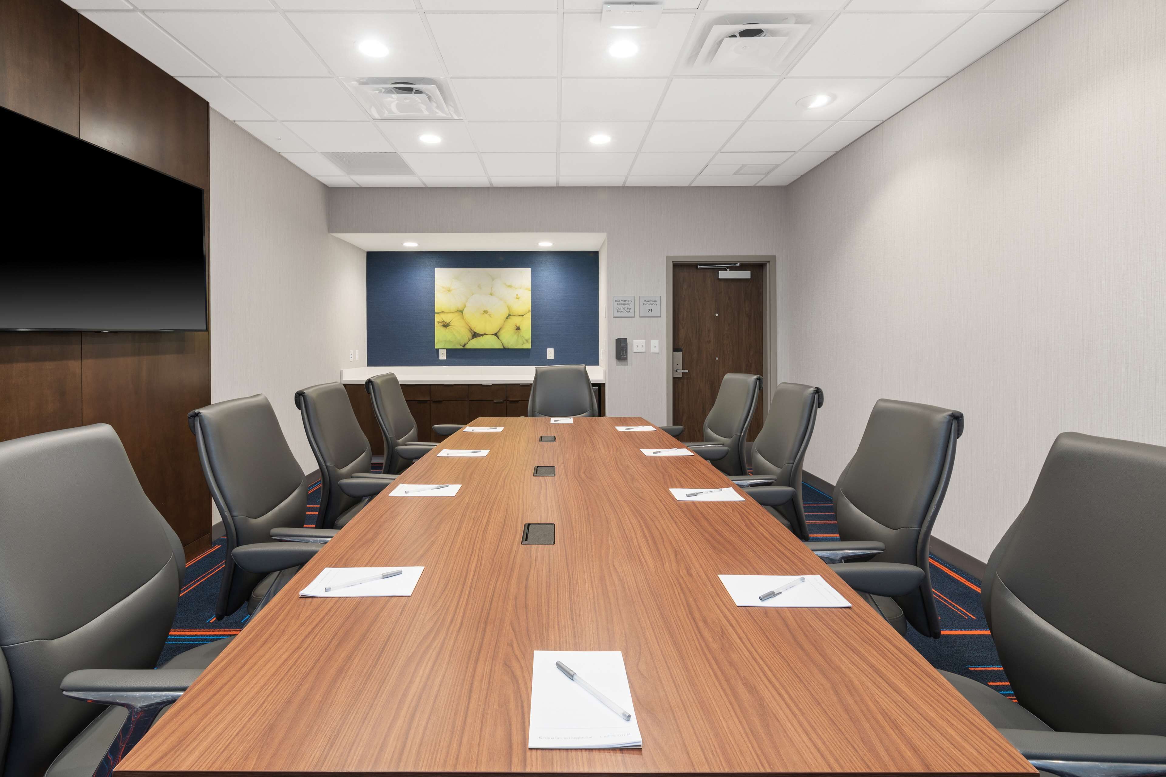 A conference room with a long wooden table, six chairs, and a large flat-screen TV on the wall.