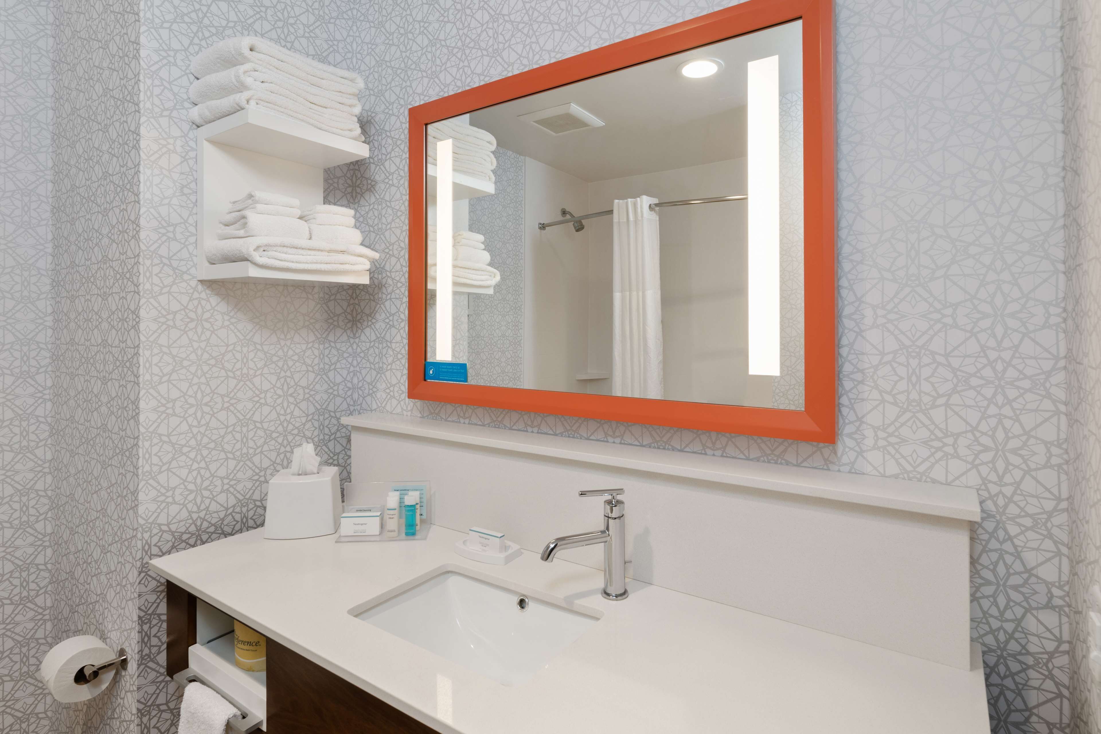 A bathroom with a white sink, a mirror with an orange frame, and shelves with folded towels.