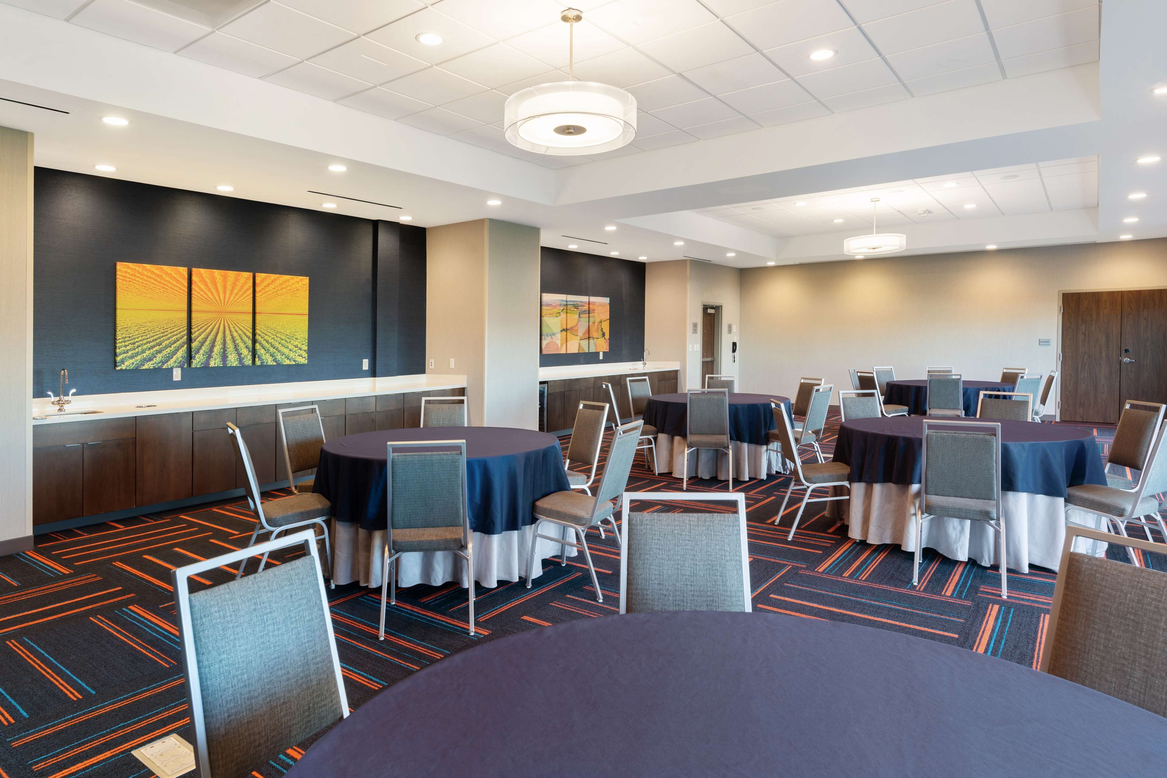 Une salle de conférence avec des tables et des chaises rondes, un grand tableau accroché au mur et un sol recouvert de moquette.