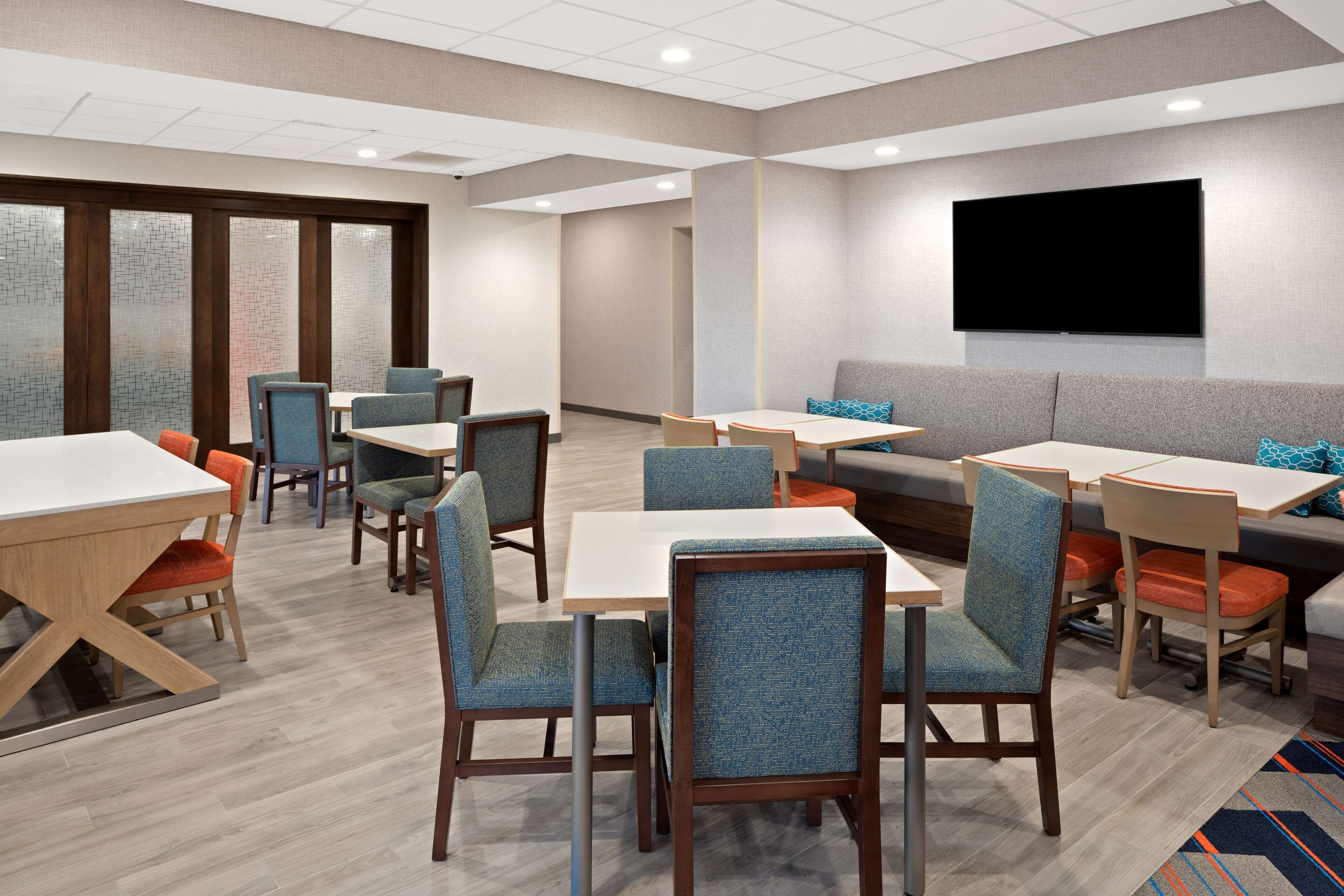 A hotel lobby with multiple seating areas, including tables and chairs, a large flat-screen TV mounted on the wall, and a patterned floor.
