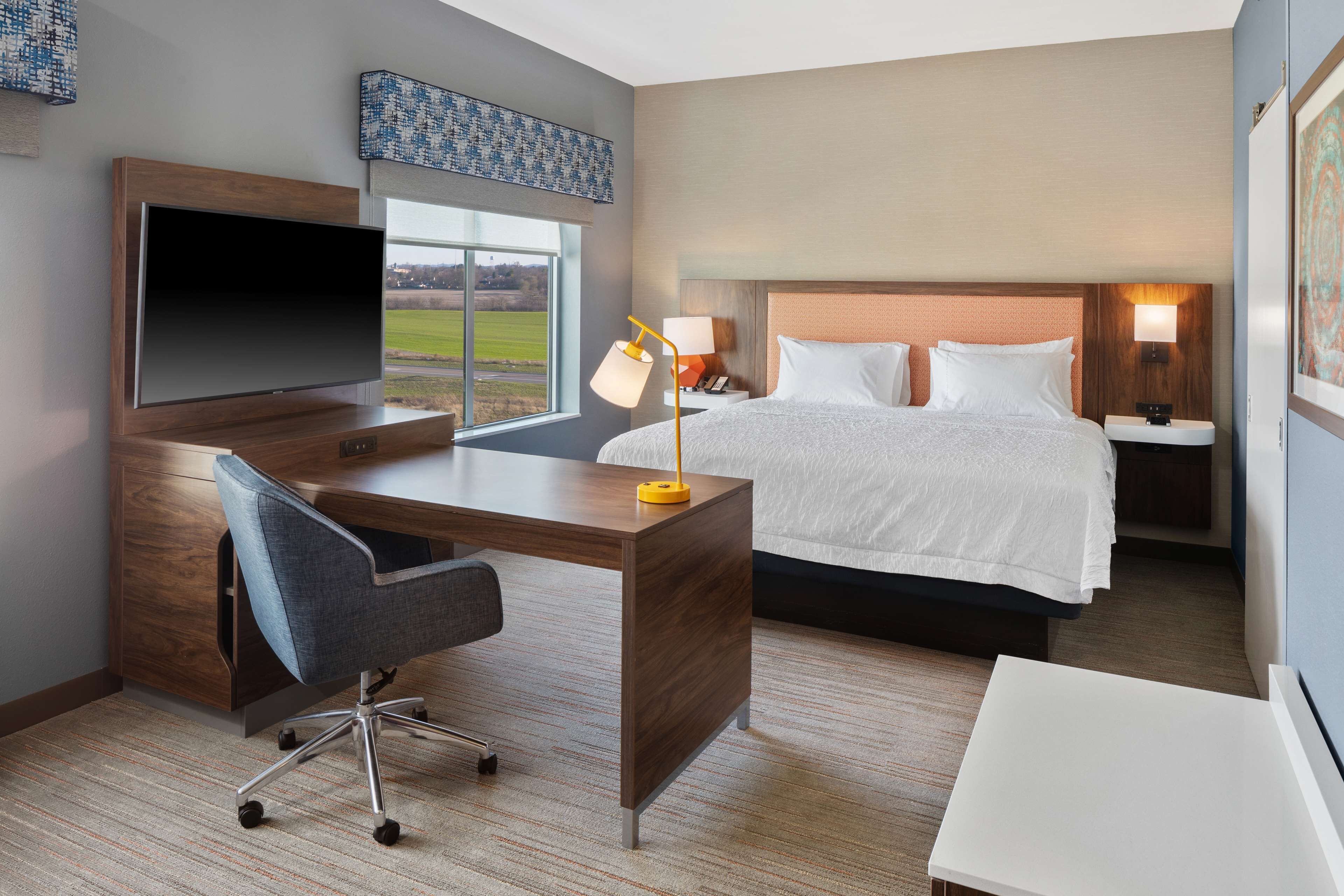A hotel room with a large bed, a desk with a TV, a chair, and a bedside table with a lamp.