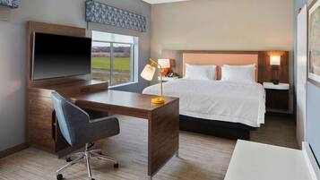 A hotel room with a large bed, a desk with a TV, a chair, and a bedside table with a lamp.