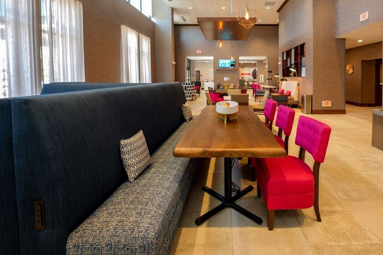 A modern hotel lobby with a long wooden table, a black sofa with patterned cushions, and bright red chairs.