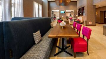 A modern hotel lobby with a long wooden table, a black sofa with patterned cushions, and bright red chairs.
