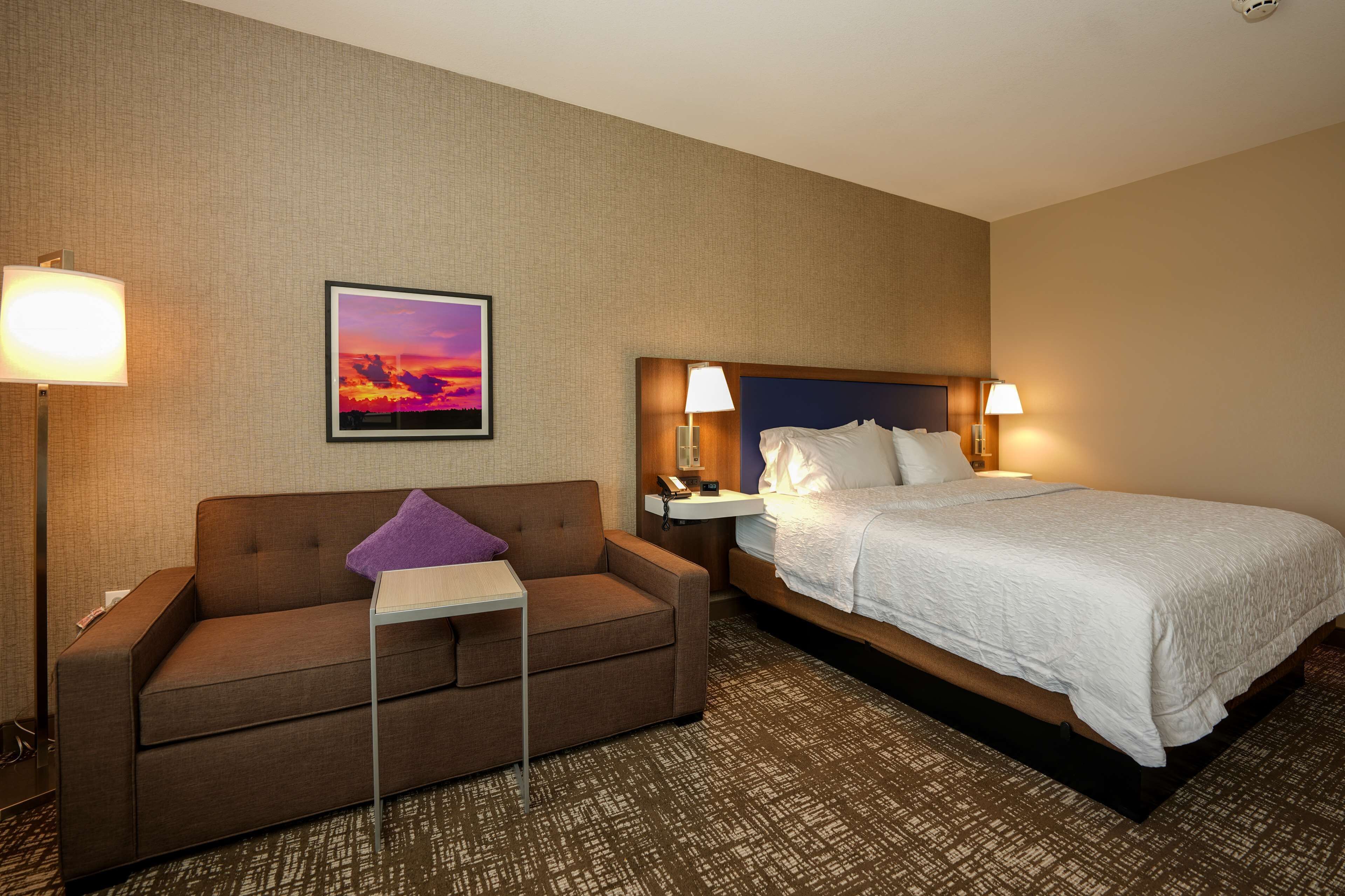 Habitación de hotel con cama, sofá, mesita de noche, lámpara y un cuadro en la pared.
