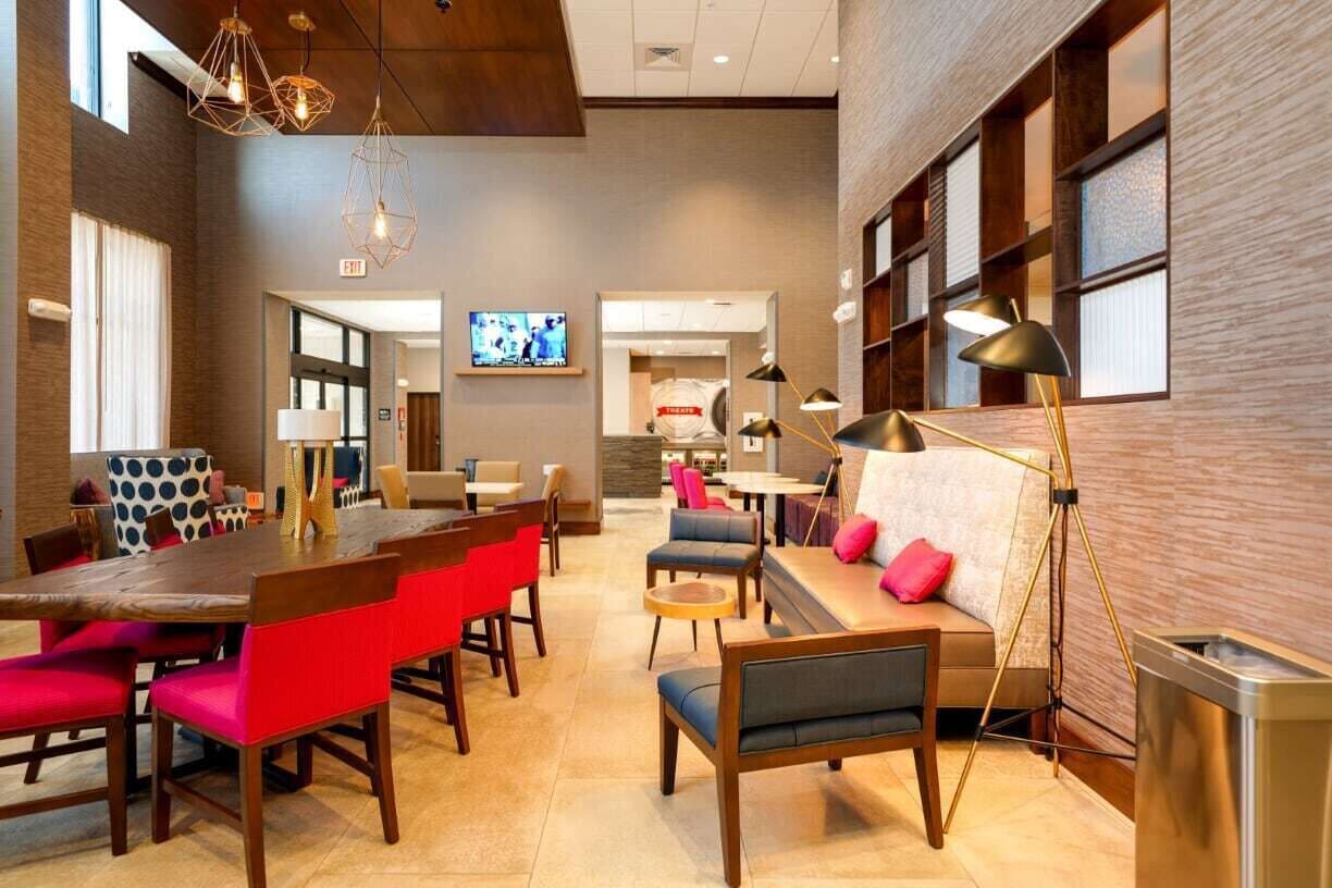 A modern hotel lobby with a wooden reception desk, red and blue seating, and a TV screen in the background.