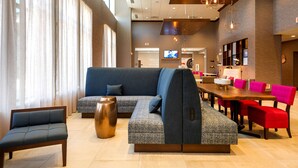 Eine moderne Hotelhalle mit einer großen Eckcouch, einem Essbereich mit roten Stühlen und einem Bildschirm, der eine Willkommensbotschaft zeigt.