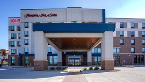 Eingangsbereich eines Hampton Inn Suites Hotels mit modernem Design, gekennzeichnet durch einen markanten Eingang, große Fenster und einen wolkenlosen Himmel.