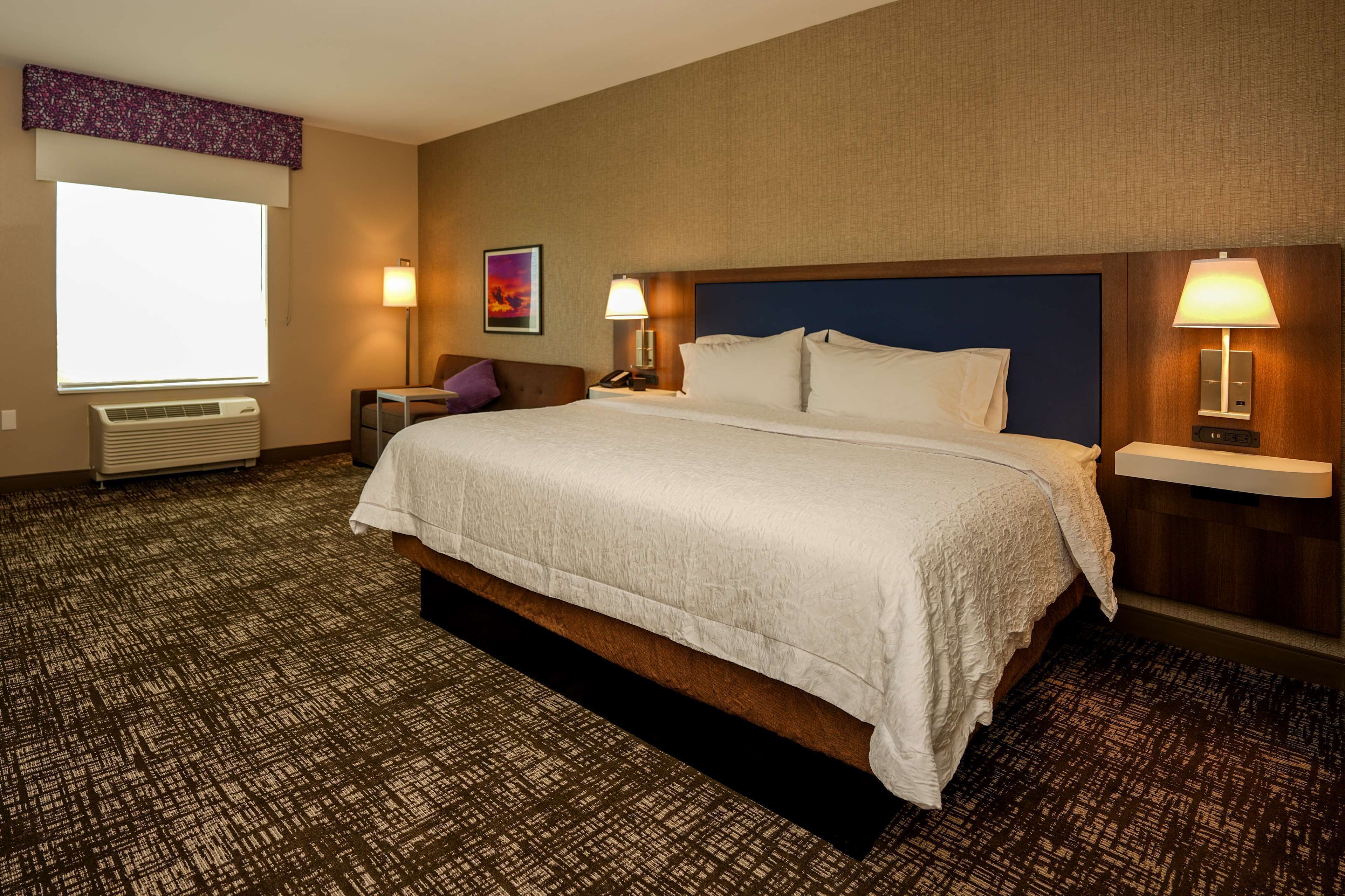 A hotel room with a large bed, two bedside tables with lamps, a chair, a desk, and a window with a view.