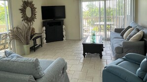 A living room with a ceiling fan, a television, two sofas, a coffee table, and a large window with blinds. - Little Gasparilla Island Getaway (Placida)