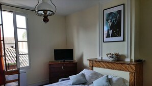 A bedroom with a bed, a television on a wooden dresser, a framed artwork on the wall, and a window with a view of buildings. - Large charming studio (Cordes-sur-Ciel)