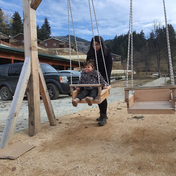 A wooden swing set in a park with a building in the background. - Danzot Tatil Köyü (Ardanuç)