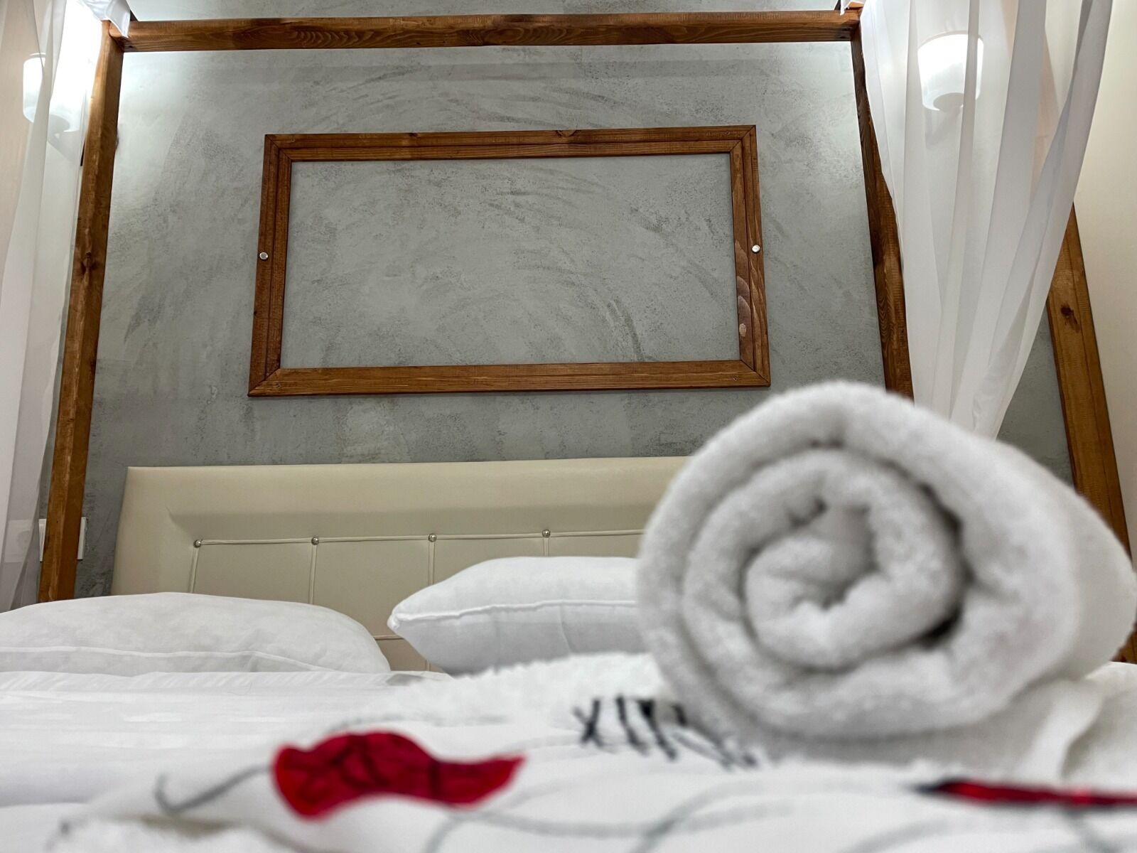 A neatly made bed with white bedding and a rolled white towel, a wooden picture frame on the wall, and a white curtain partially visible.