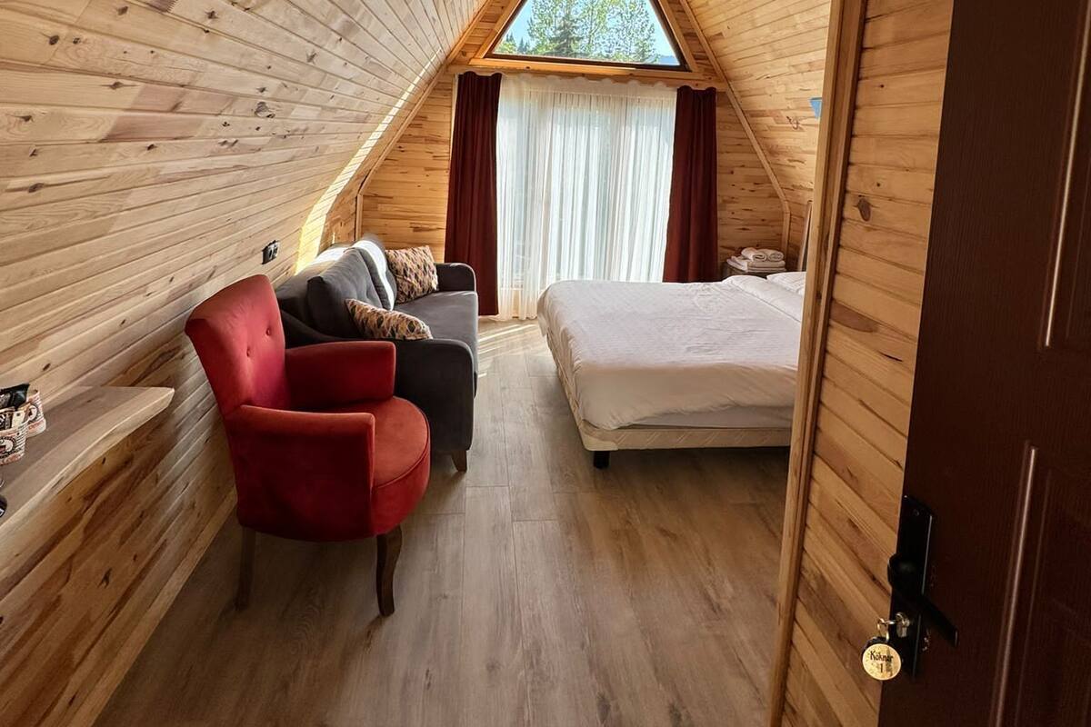 A cozy attic bedroom with wooden walls, a red chair, a bed, and a window with curtains.