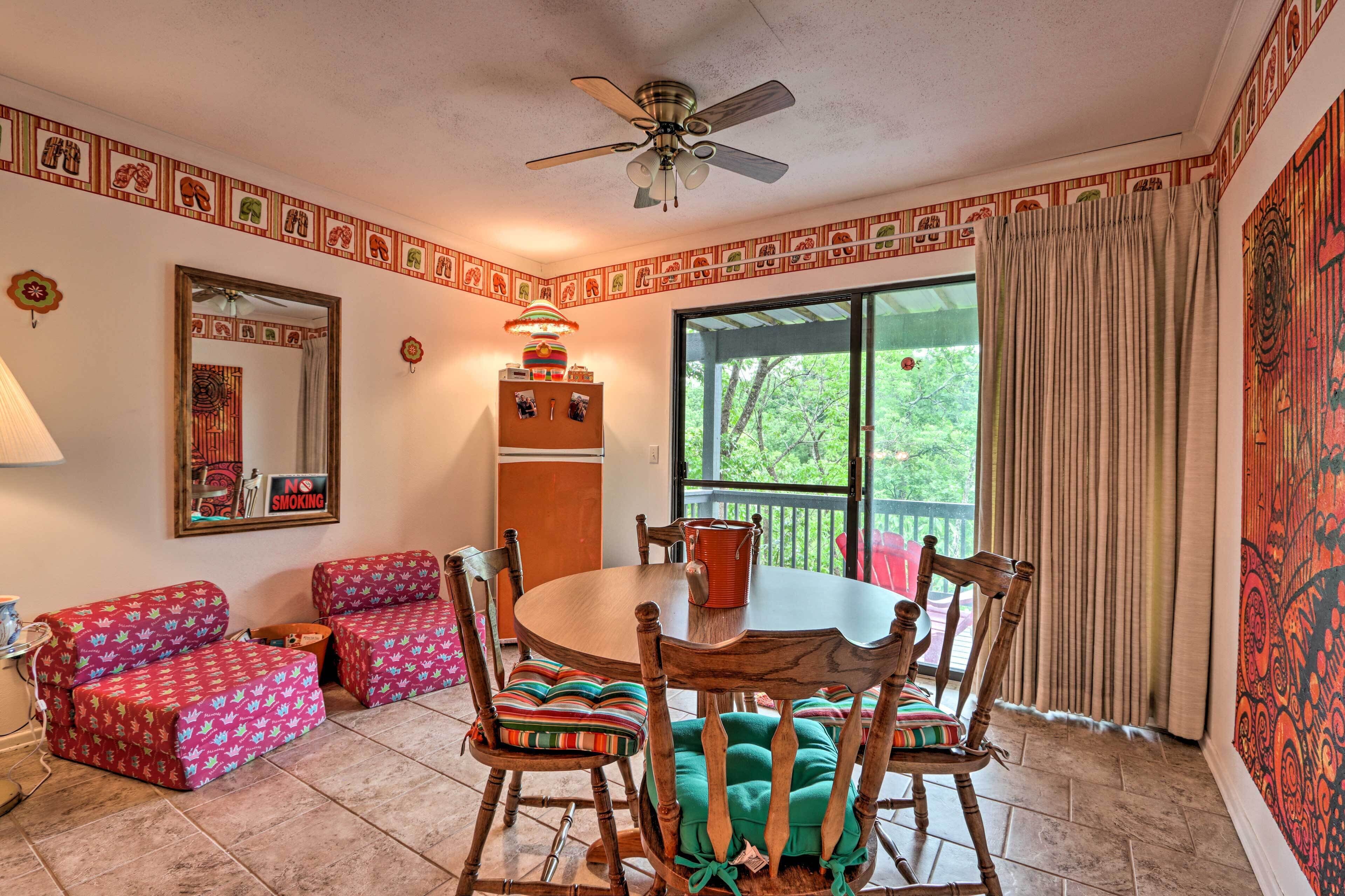 A dining area with a round table, chairs, and a ceiling fan. There is a window with curtains and a view of greenery outside.